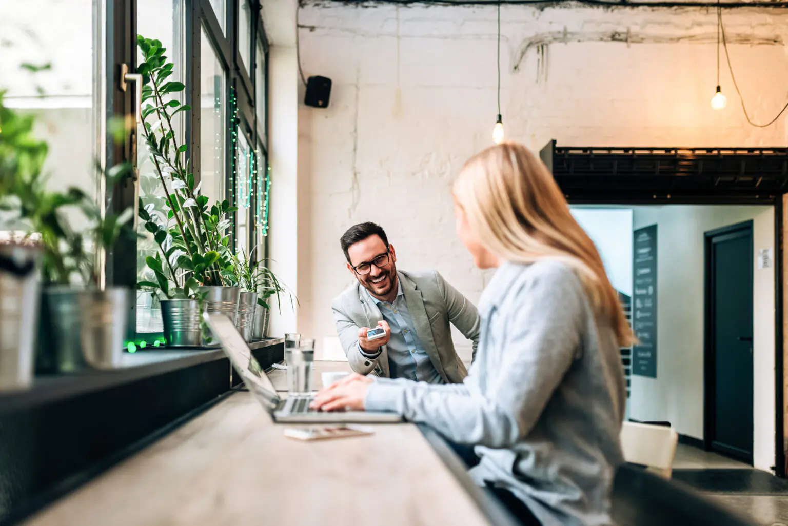 Deux professionnels échangeant dans un espace de coworking lumineux ; un homme souriant tend sa carte à une partenaire assise devant un ordinateur