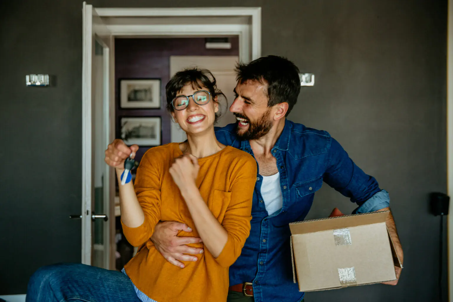 Couple heureux entrant dans leur nouveau logement avec clé et carton en main.