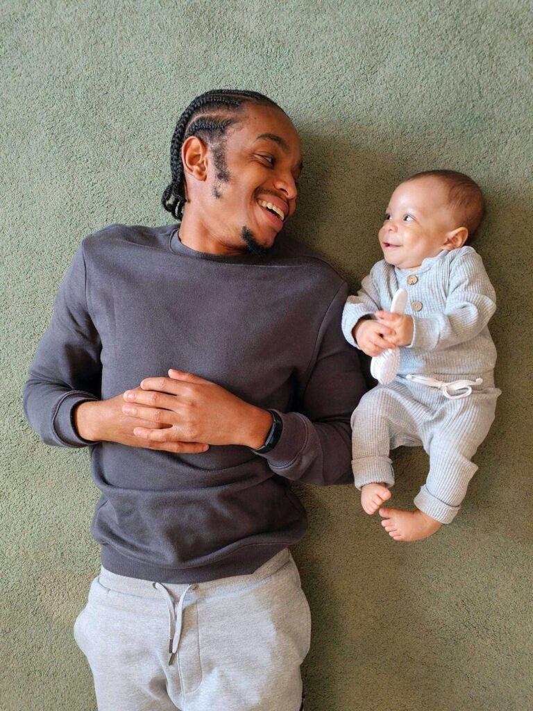 Père souriant allongé à côté de son bébé, symbolisant la protection familiale offerte par Garance